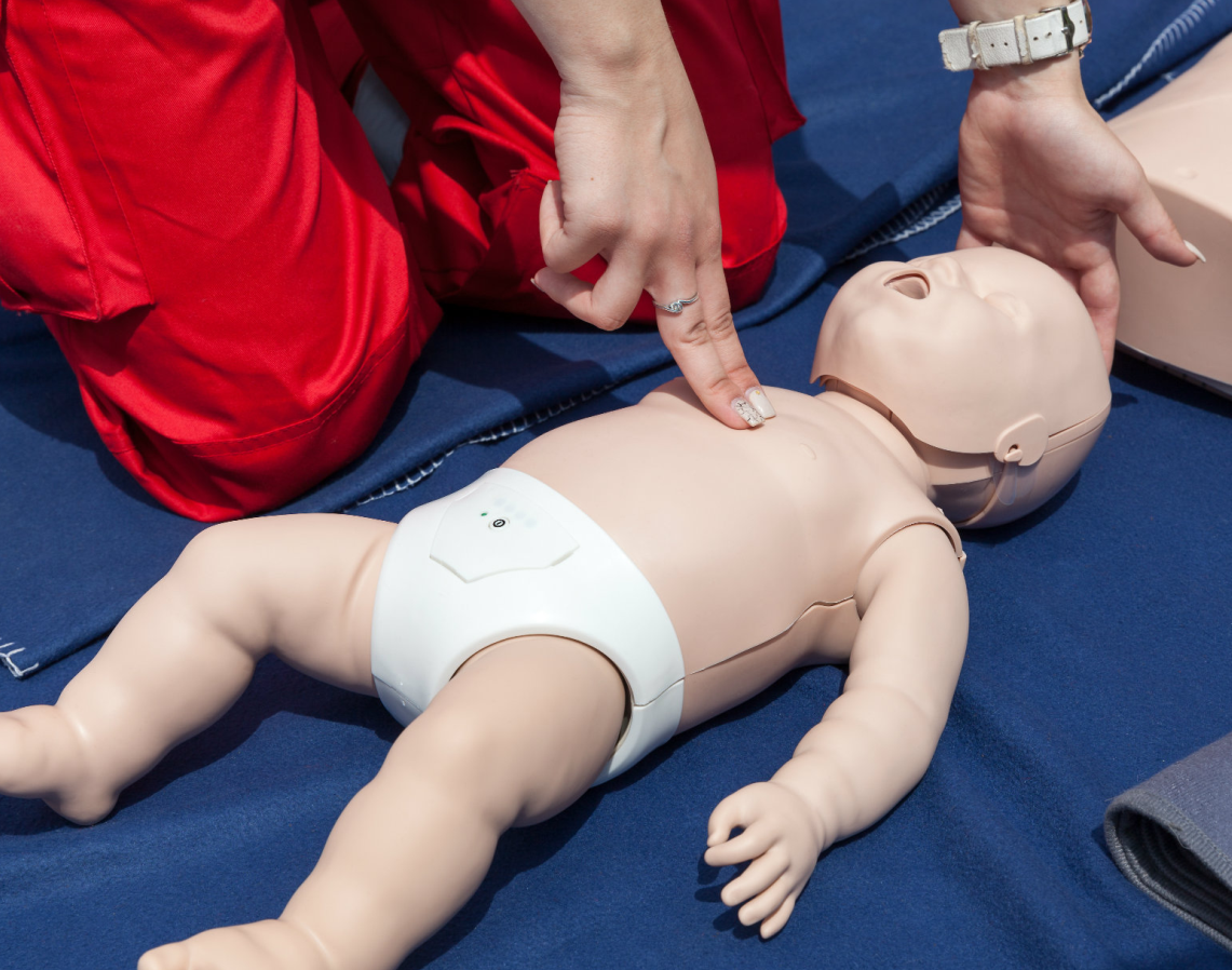 Erste Hilfe am Kind - Helferin neben einer Babywiederbelebungspuppe die mit der einen Hand den Kopf hält und mit der anderen Hand mit zwei Fingern Herzdruckmassage durchführt